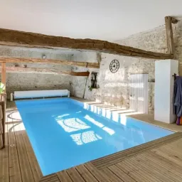 Kryty basen w rustykalnym stylu z drewnianym wykończeniem, kamiennymi ścianami i widocznymi drewnianymi belkami stropowymi, oświetlony naturalnym światłem odbijającym się w wodzie.