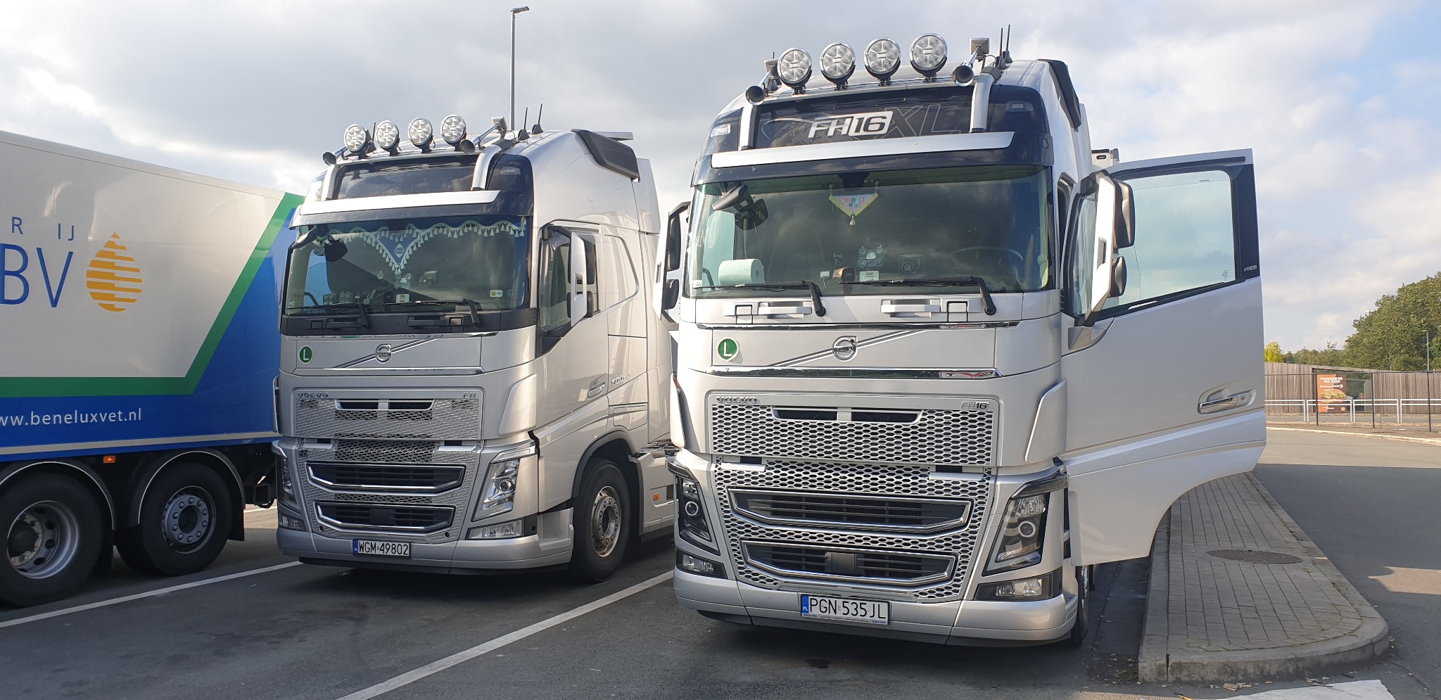 Dwa srebrne ciągniki siodłowe Volvo FH, jeden z otwartymi drzwiami, zaparkowane na parkingu, widoczny fragment naczepy chłodniczej z logo firmy.