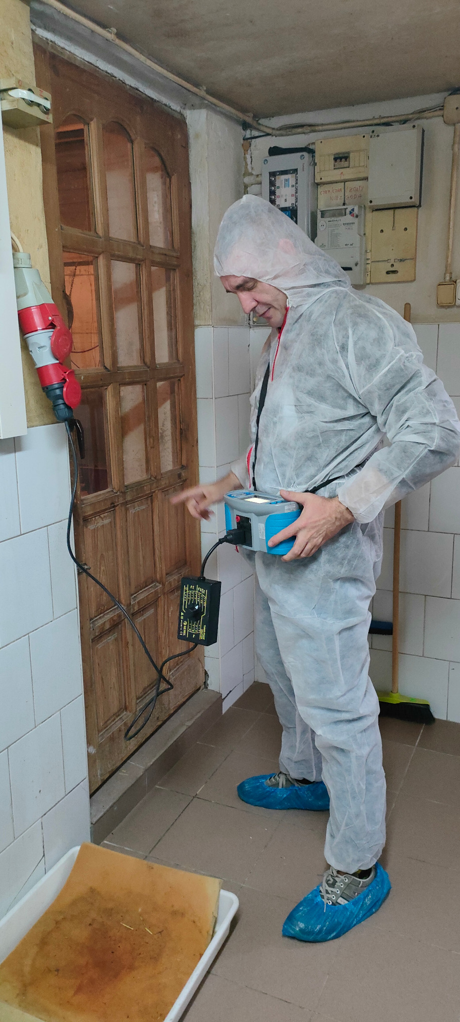 Elektryk w kombinezonie ochronnym i niebieskich ochraniaczach na obuwiu, mierzy parametry instalacji elektrycznej przy użyciu specjalistycznego urządzenia, podłączonego do gniazda siłowego.