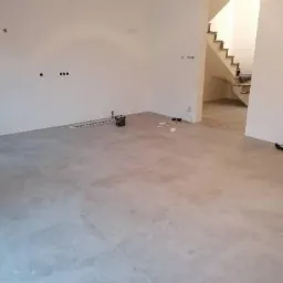 Ułożona szara podłoga z płytek wielkoformatowych w pustym pomieszczeniu z widocznymi instalacjami elektrycznymi i schodami w tle.