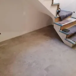 Betonowa posadzka z widocznymi śladami szlifowania, schody drewniane z zabezpieczeniem na czas remontu, surowe ściany przygotowane do dalszych prac wykończeniowych.