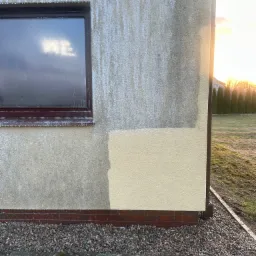 Fragment elewacji budynku z widocznym oknem i próbką nowej farby, na tle zachodzącego słońca i zieleni.