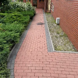 Czerwona kostka brukowa tworzy ścieżkę obok zieleni i ceglanej ściany budynku. Widoczny manhole. Szara obwódka kostki. Ujęcie z perspektywy pieszego, słoneczny dzień.