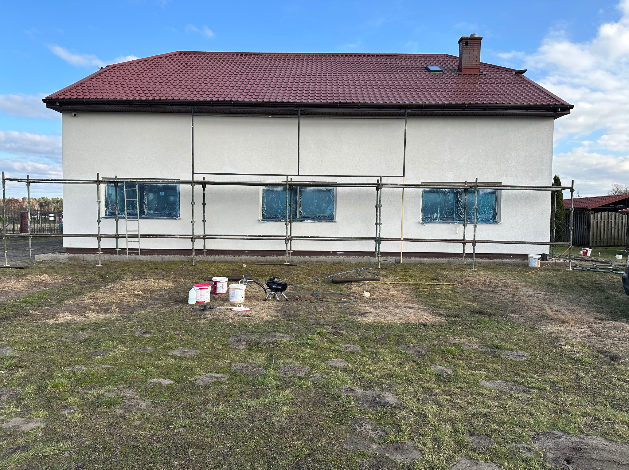 Przygotowania do prac elewacyjnych w Sianowie: dom z zabezpieczonymi oknami, rusztowanie, wiaderka z farbą i agregat malarski na trawniku.