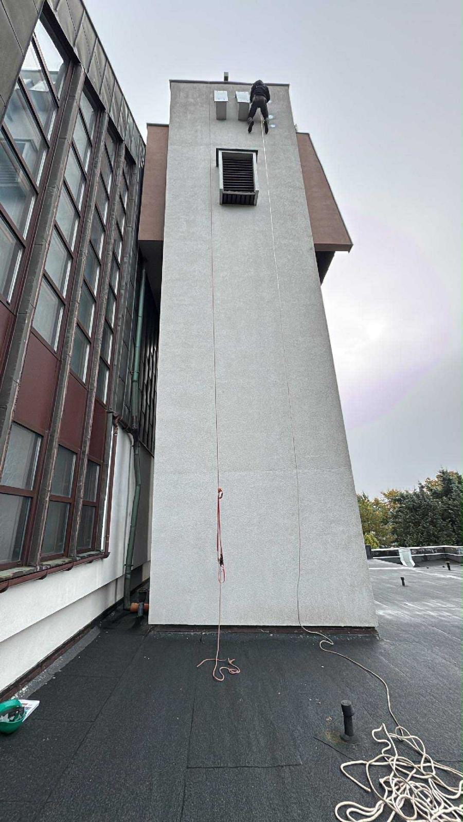 Pracownik na linie, alpinistycznie, na tle wysokiej wieży budynku z szarą elewacją. Widok z dachu, liny i sprzęt asekuracyjny. Praca na wysokościach.