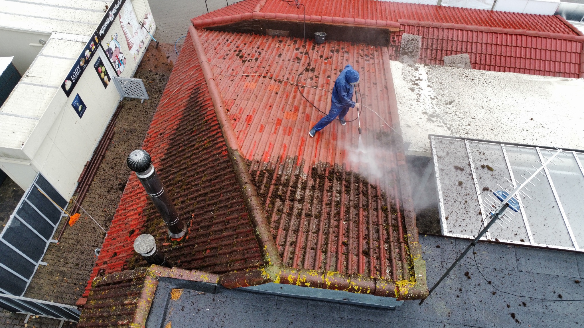 Czerwony dach z blachy falistej, częściowo wyczyszczony z mchu i zabrudzeń przez osobę w niebieskim kombinezonie za pomocą myjki ciśnieniowej. Widok z góry.