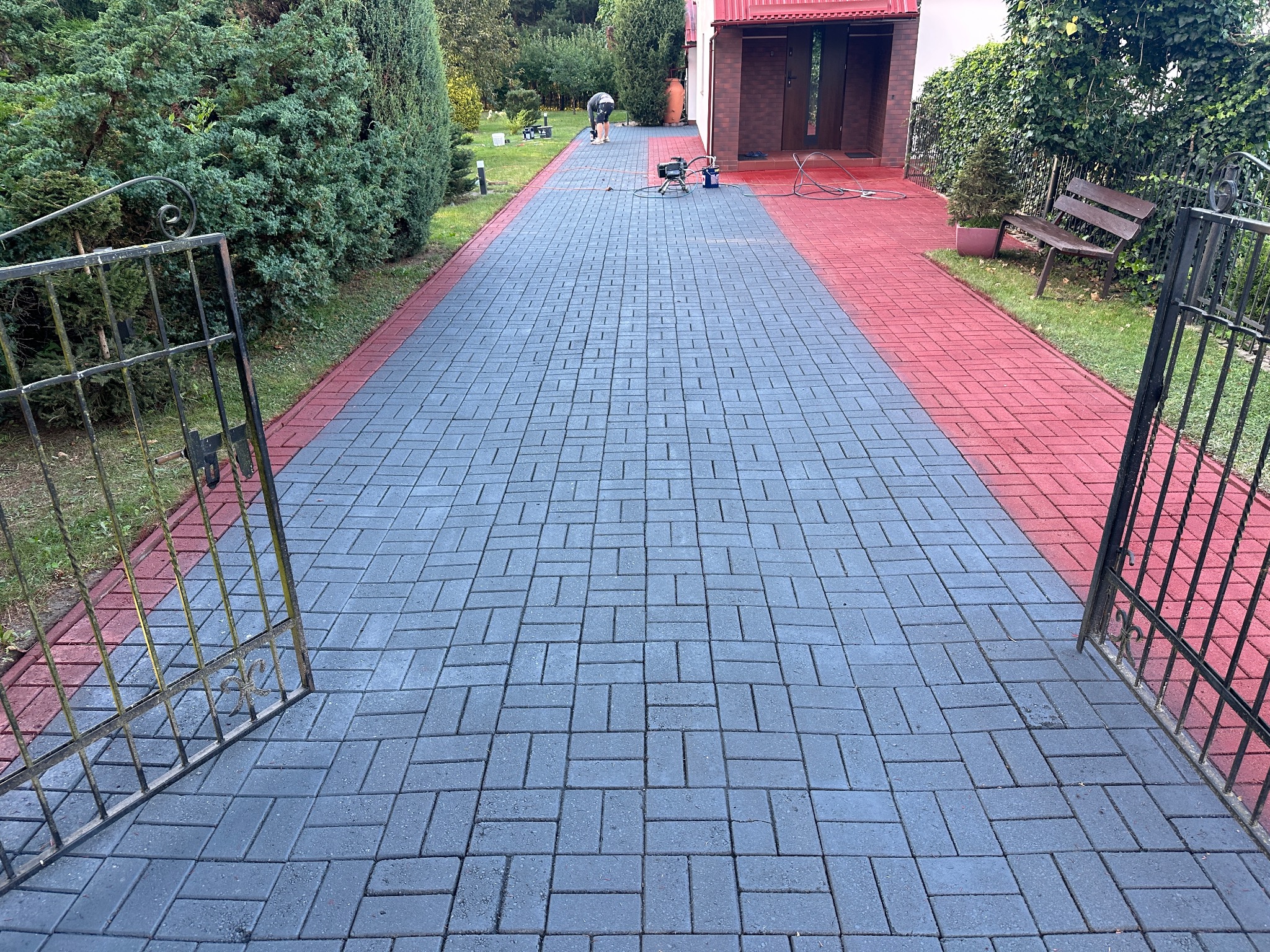 Malowanie kostki brukowej na posesji. Część kostki pomalowana na czerwono, reszta szara. Widoczny sprzęt do malowania i osoba wykonująca pracę. Otwarta brama wjazdowa.