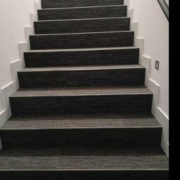 Artino Podłogi - Schody pokryte ciemną wykładziną dywanową z metalowymi listwami na krawędziach stopni, oświetlenie punktowe na ścianie.