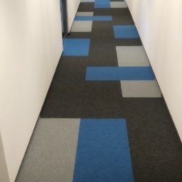 Artino Podłogi - Długi korytarz biurowy z wykładziną dywanową w geometryczny wzór z kwadratów w kolorach: czarnym, szarym i niebieskim. Po prawej stronie widoczna gaśnica.