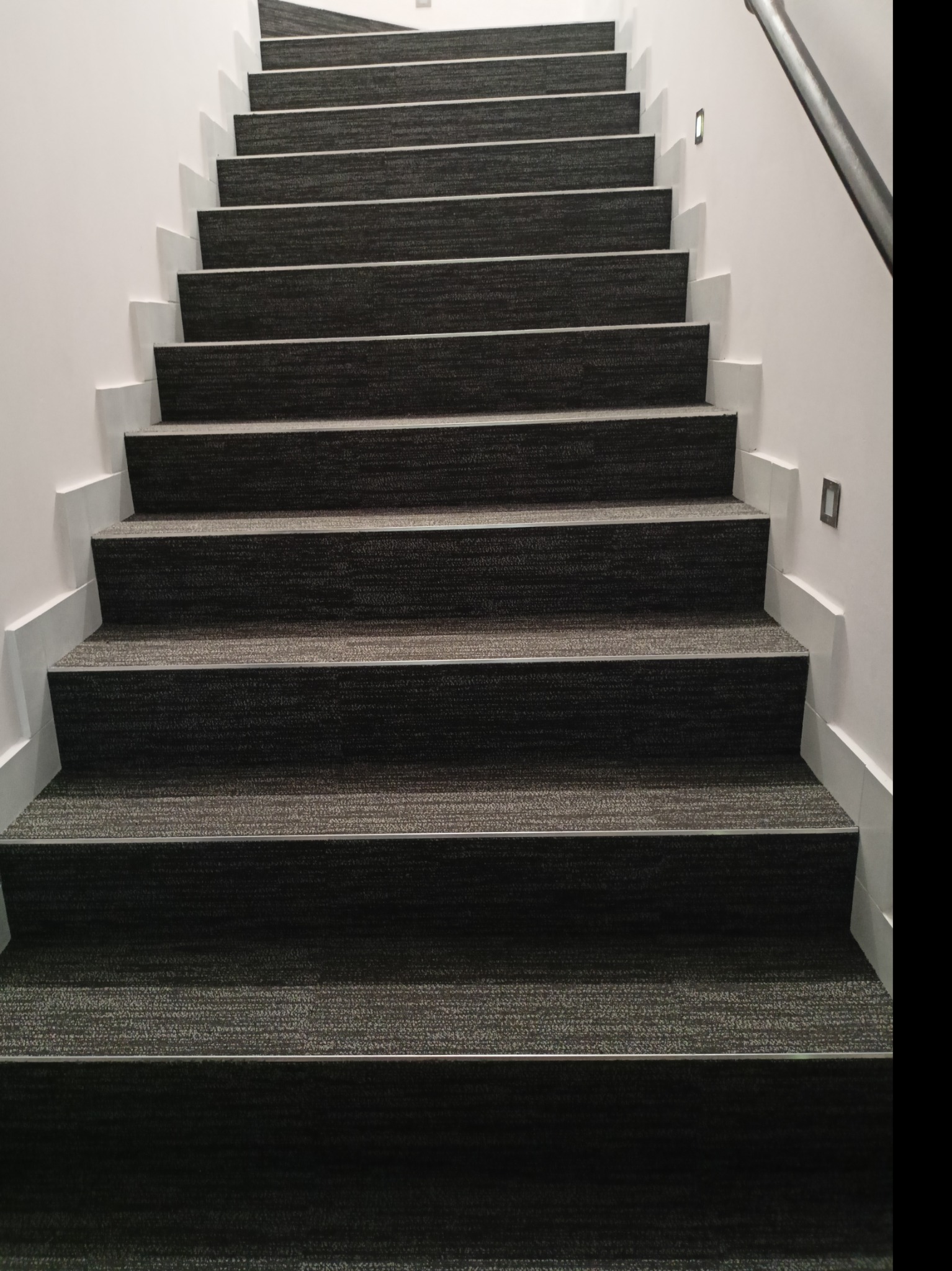 Schody pokryte ciemną wykładziną dywanową z metalowymi listwami na krawędziach stopni, oświetlenie punktowe na ścianie.