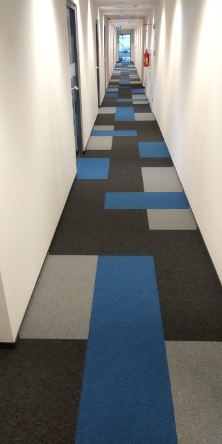 Długi korytarz biurowy z wykładziną dywanową w geometryczny wzór z kwadratów w kolorach: czarnym, szarym i niebieskim. Po prawej stronie widoczna gaśnica.
