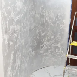 Ściana pokryta dekoracyjną, srebrzystą fakturą z efektem chmur, widoczna drabina aluminiowa i biała marmurowa podłoga.