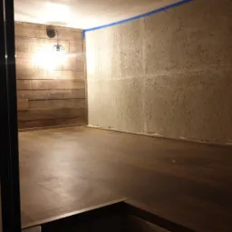 Wnętrze pokoju z drewnianą boazerią na jednej ścianie i szpachlowaną, surową ścianą z niebieską taśmą malarską na górze, drewniana podłoga, widok przez metalowe pręty.