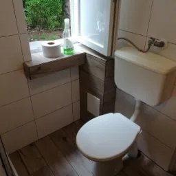 Toaleta kompaktowa z białą ceramiką, zamontowana w wykończonej łazience z jasnymi płytkami ściennymi i podłogą imitującą drewno; na półce przy oknie papier toaletowy i butelka płynu do czyszczenia.