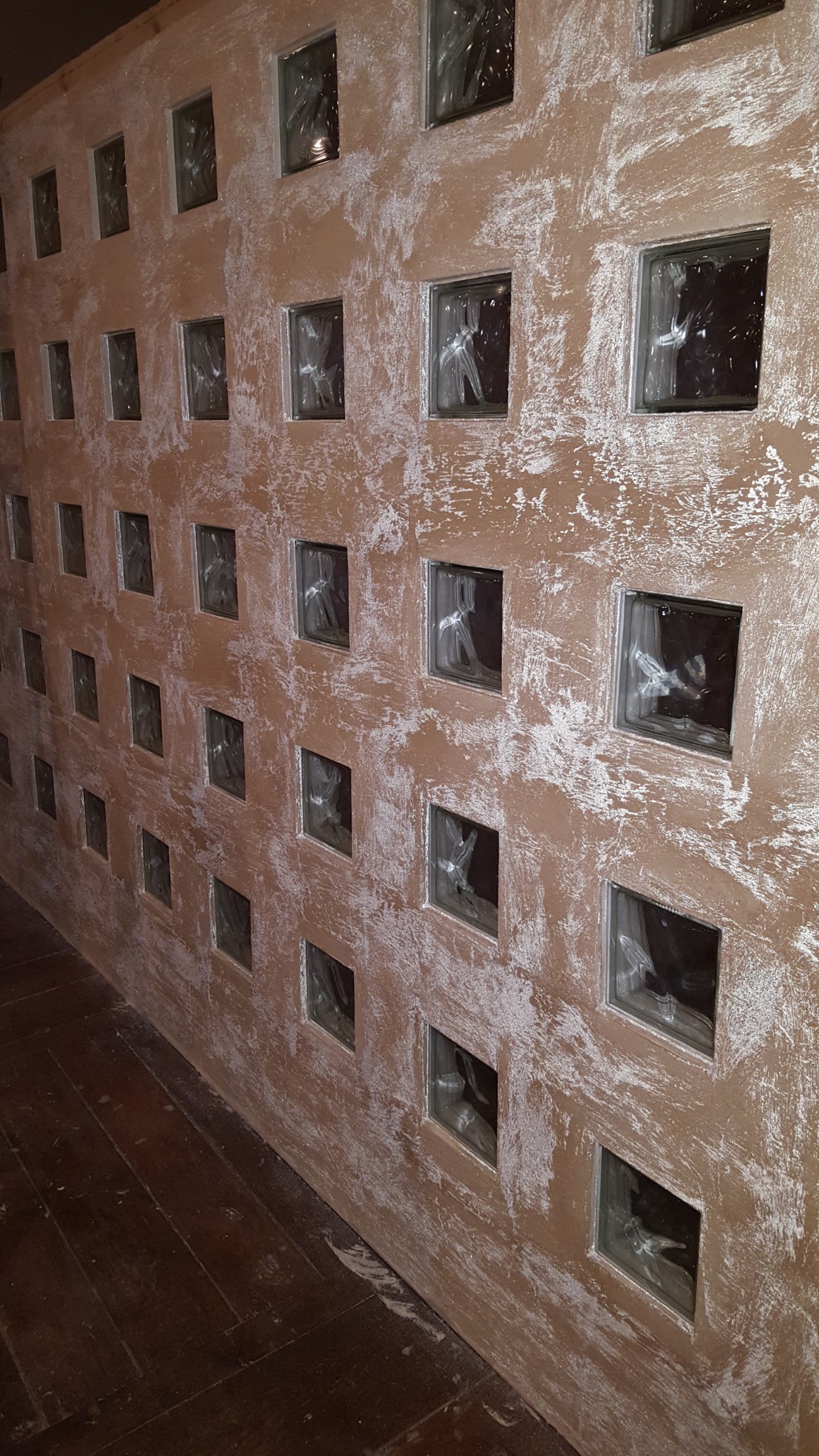 Ściana z dekoracyjnym szpachlowaniem w odcieniach beżu i bieli, z wkomponowanymi szklanymi luksferami, widok z perspektywy podłogi z ciemnego drewna.