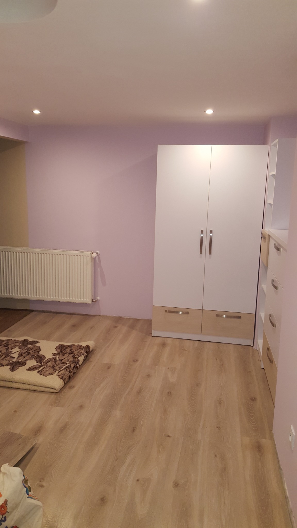 Pokój po odświeżeniu: Ściany pomalowane na fioletowo, jasna podłoga imitująca drewno, biała szafa z drewnianymi akcentami, grzejnik na ścianie i wzorzysty dywanik na podłodze.