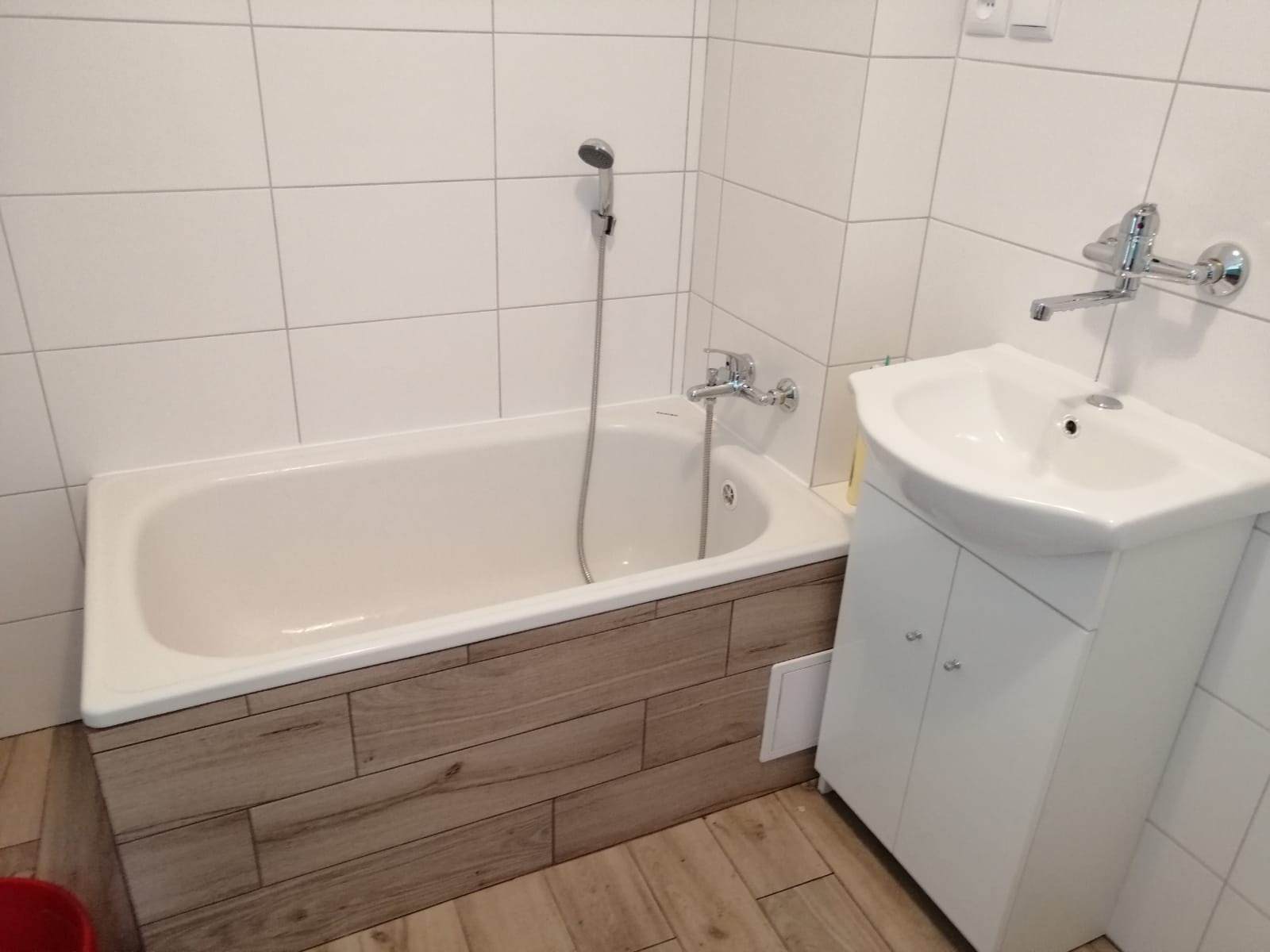 Biała wanna obudowana panelami imitującymi drewno, biała umywalka z szafką, chromowana bateria i słuchawka prysznicowa na tle białych płytek.