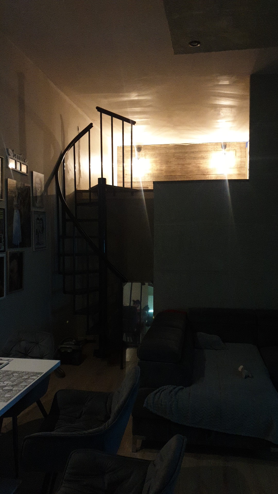 Wnętrze mieszkania z spiralnymi schodami prowadzącymi na antresolę, oświetloną drewnianą ścianą i dwoma lampami, widoczne zdjęcia na ścianie i szare meble.