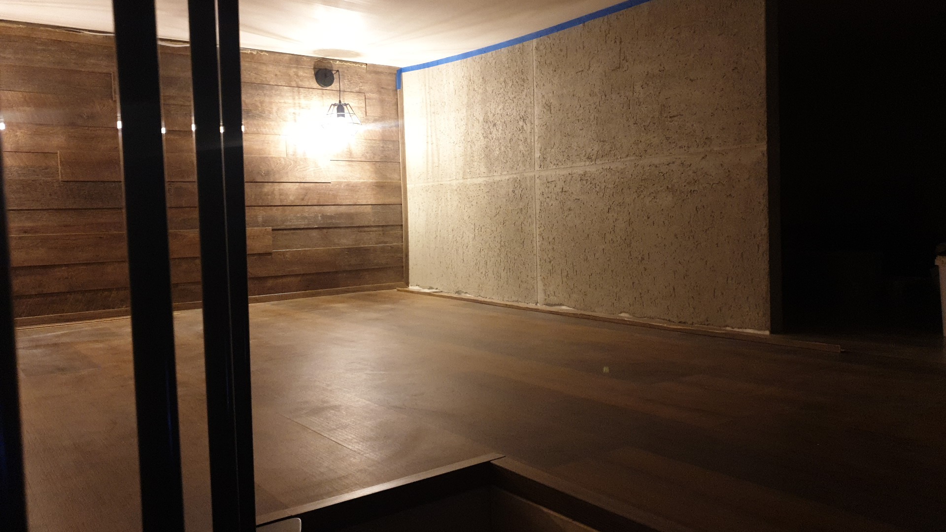 Wnętrze pokoju z drewnianą boazerią na jednej ścianie i szpachlowaną, surową ścianą z niebieską taśmą malarską na górze, drewniana podłoga, widok przez metalowe pręty.