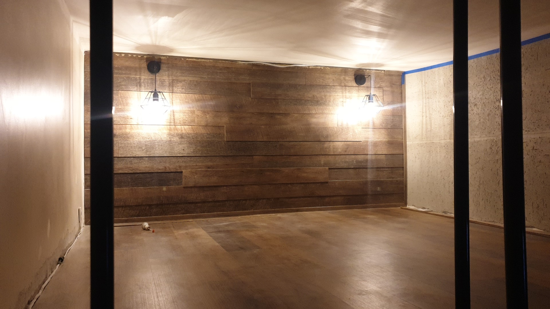 Pokój po remoncie z drewnianą ścianą, parkietem na podłodze i dwoma lampami wiszącymi, widoczne ślady po taśmie malarskiej na ścianie.