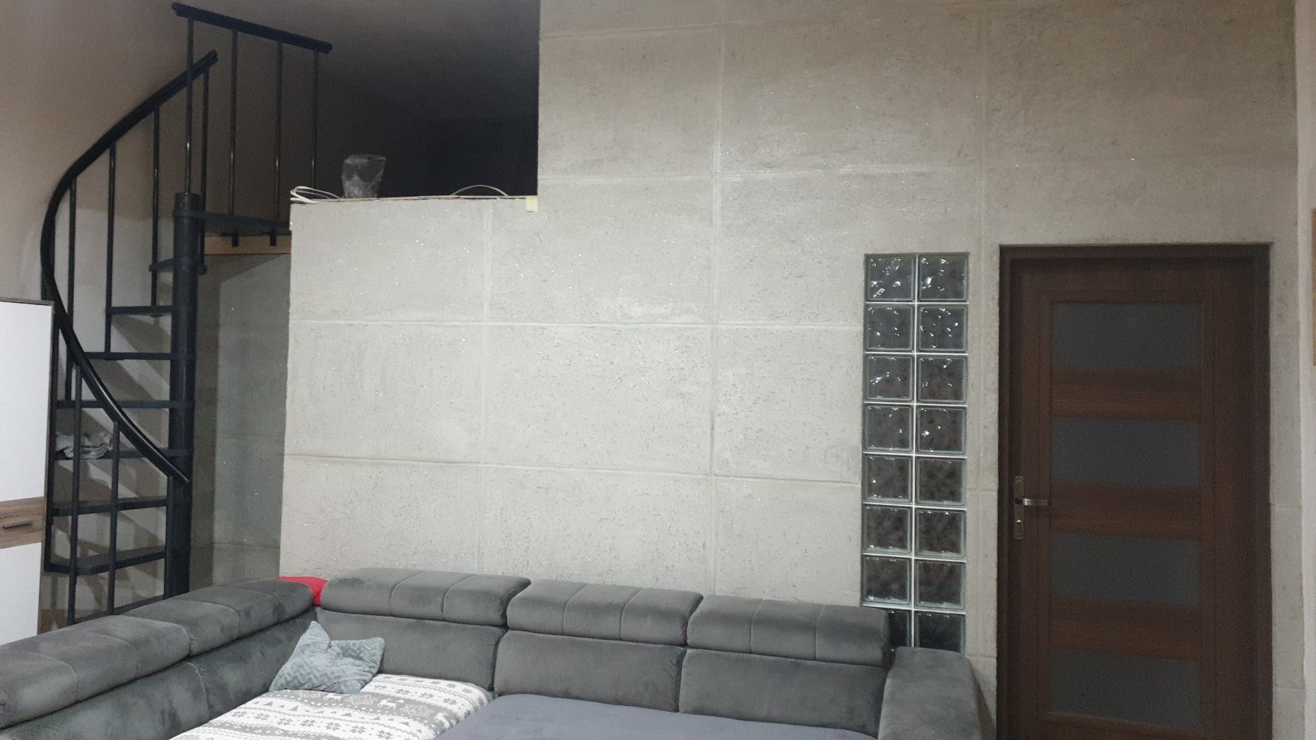 Wnętrze pokoju z spiralnymi schodami, ścianą wyłożoną dużymi, jasnymi płytami, kolumną z luksferów i ciemnymi drzwiami, widoczne fragmenty szarej sofy.