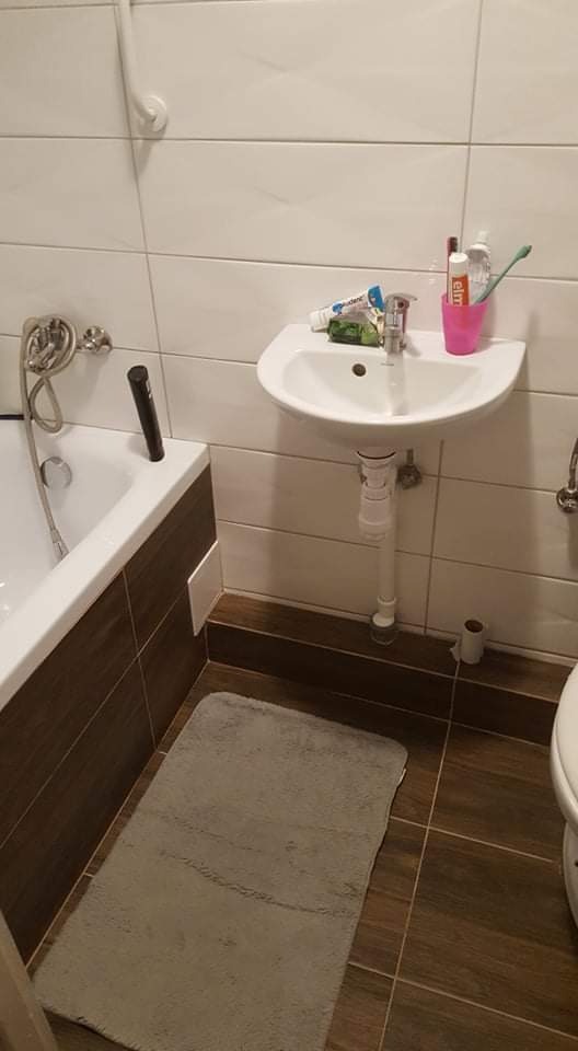 Umywalka ceramiczna zamontowana na ścianie w łazience z białymi płytkami i brązowymi akcentami, widoczne kosmetyki i dywanik podłogowy.