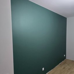 COMFORT HOME Sp. z o.o - Pomieszczenie po remoncie: ściana pomalowana na ciemnozielony kolor, jasna podłoga z paneli, widoczne gniazdka elektryczne i przełącznik światła.