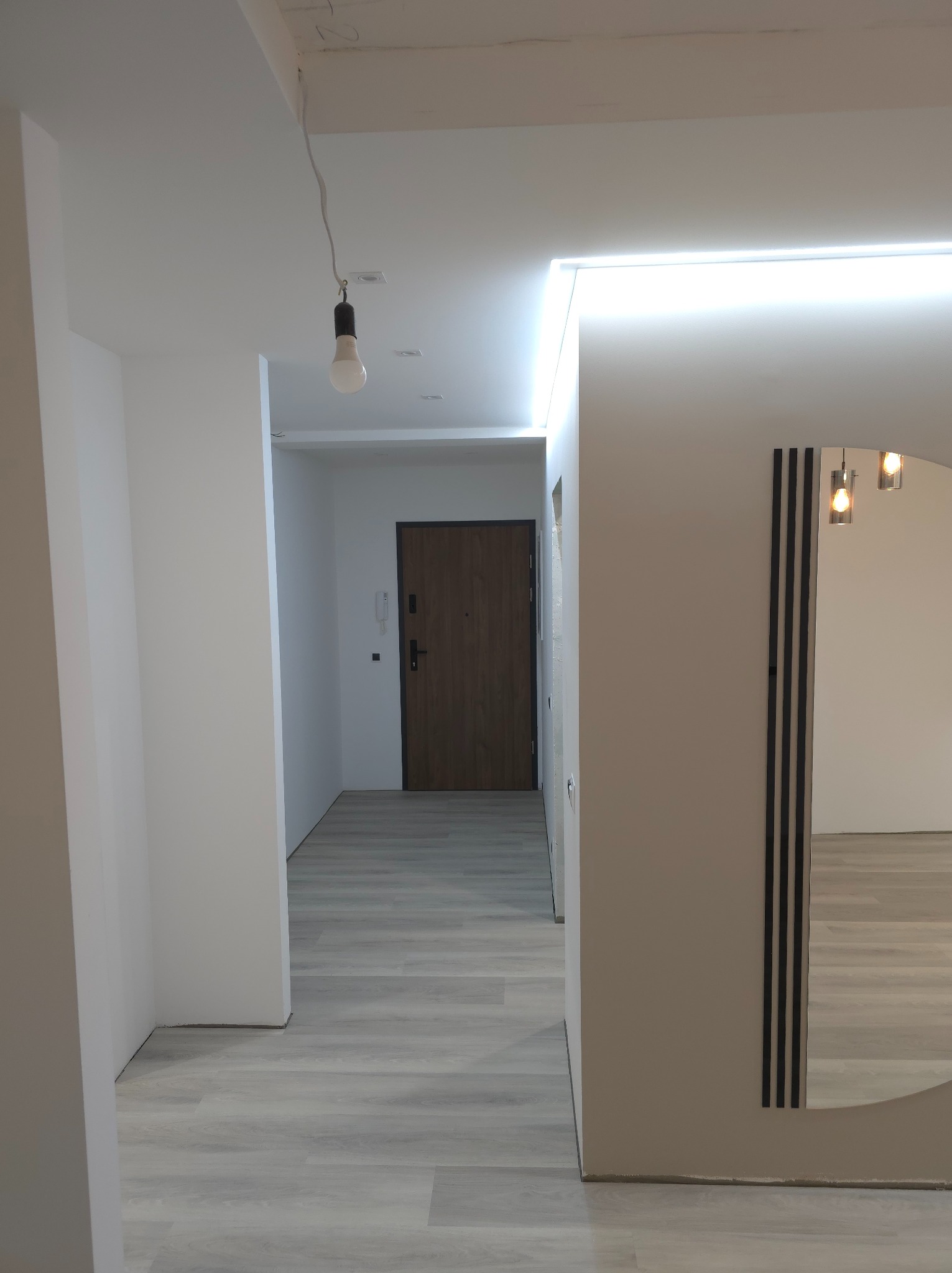 Wnętrze korytarza po remoncie: białe ściany z oświetleniem LED podsufitowym, szare panele podłogowe, drewniane drzwi wejściowe i dekoracyjne lustro z czarnymi paskami.