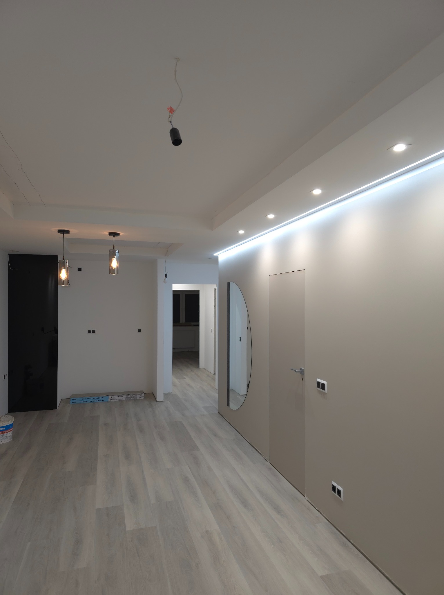 Wnętrze mieszkania w trakcie wykańczania: jasne panele podłogowe, ściany w odcieniach beżu z oświetleniem LED podsufitowym, okrągłe lustro, wiszące lampy i widoczne puszki elektryczne bez osprzętu.