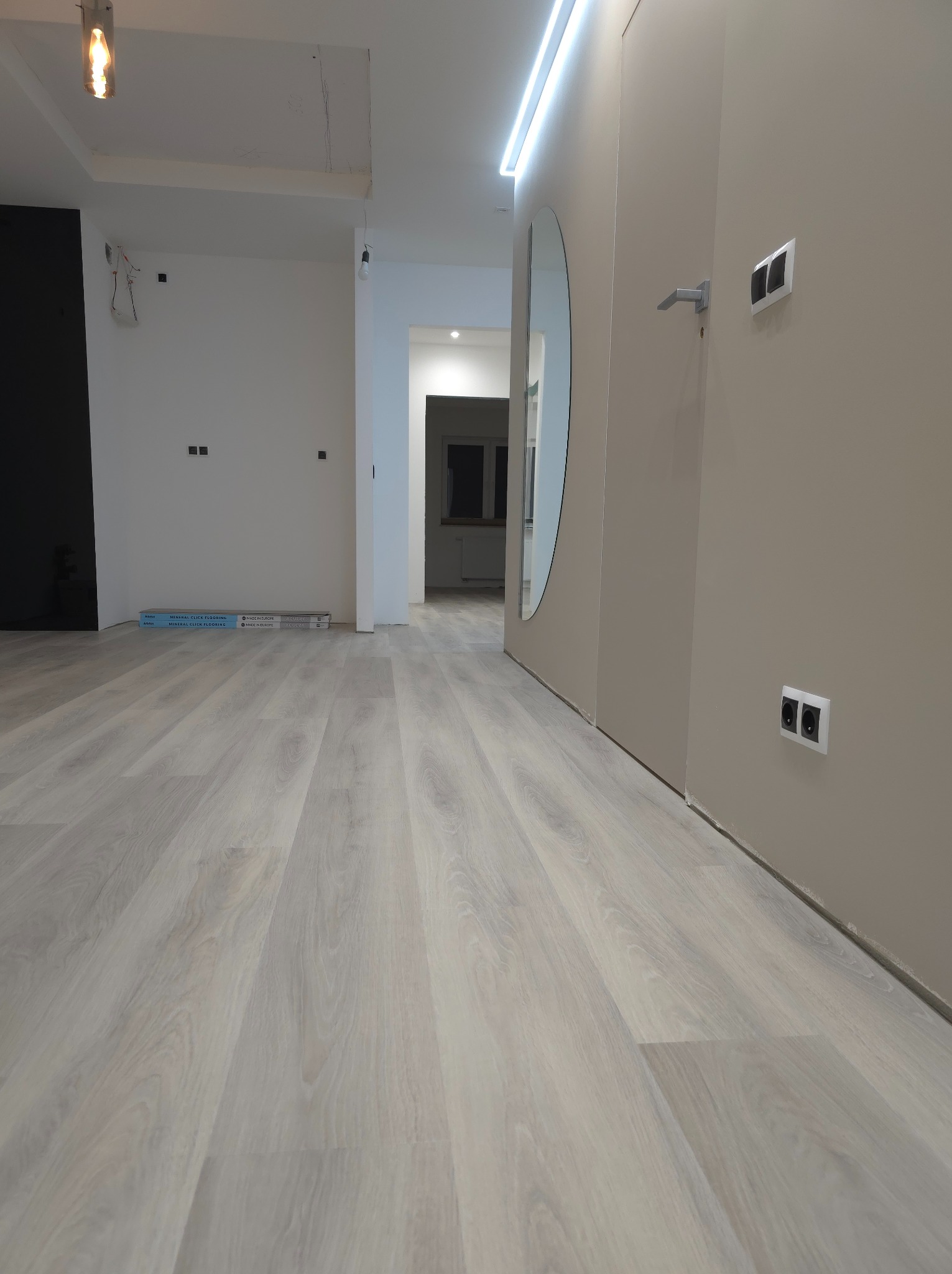 Jasne panele podłogowe ułożone w nowym mieszkaniu, widoczne paczki paneli pod ścianą, szare ściany z zamontowanymi gniazdkami elektrycznymi, w tle widok na kolejne pomieszczenie.