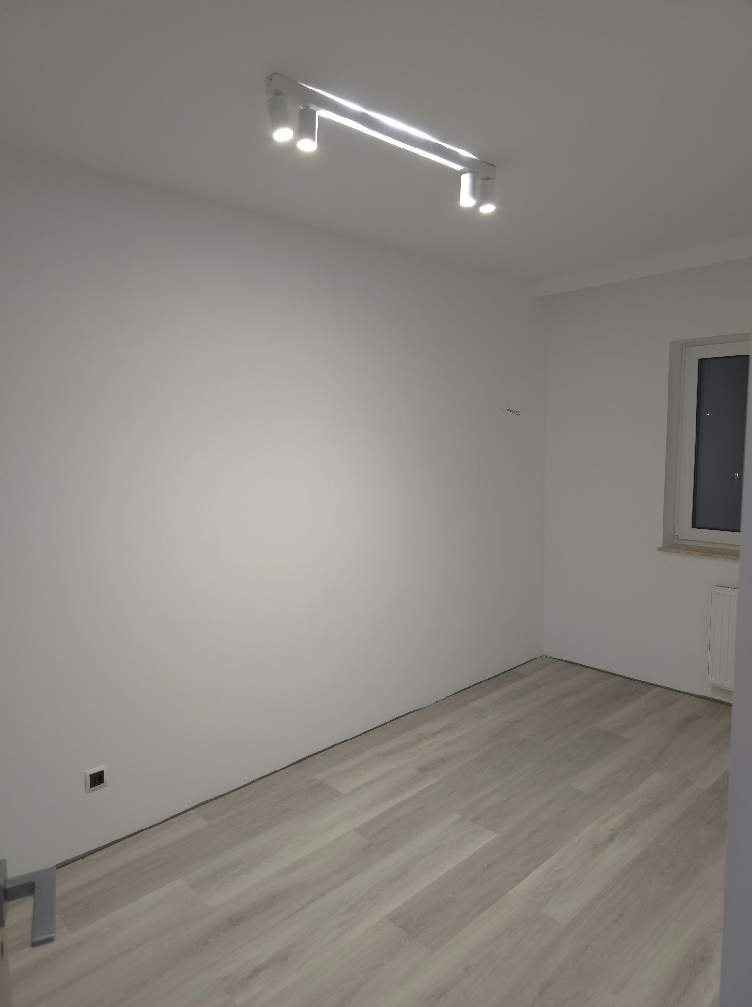 Pusty, świeżo wyremontowany pokój z panelami podłogowymi w odcieniach szarości, białymi ścianami i nowoczesnym oświetleniem sufitowym z czterema reflektorami, okno z grzejnikiem po prawej stronie.