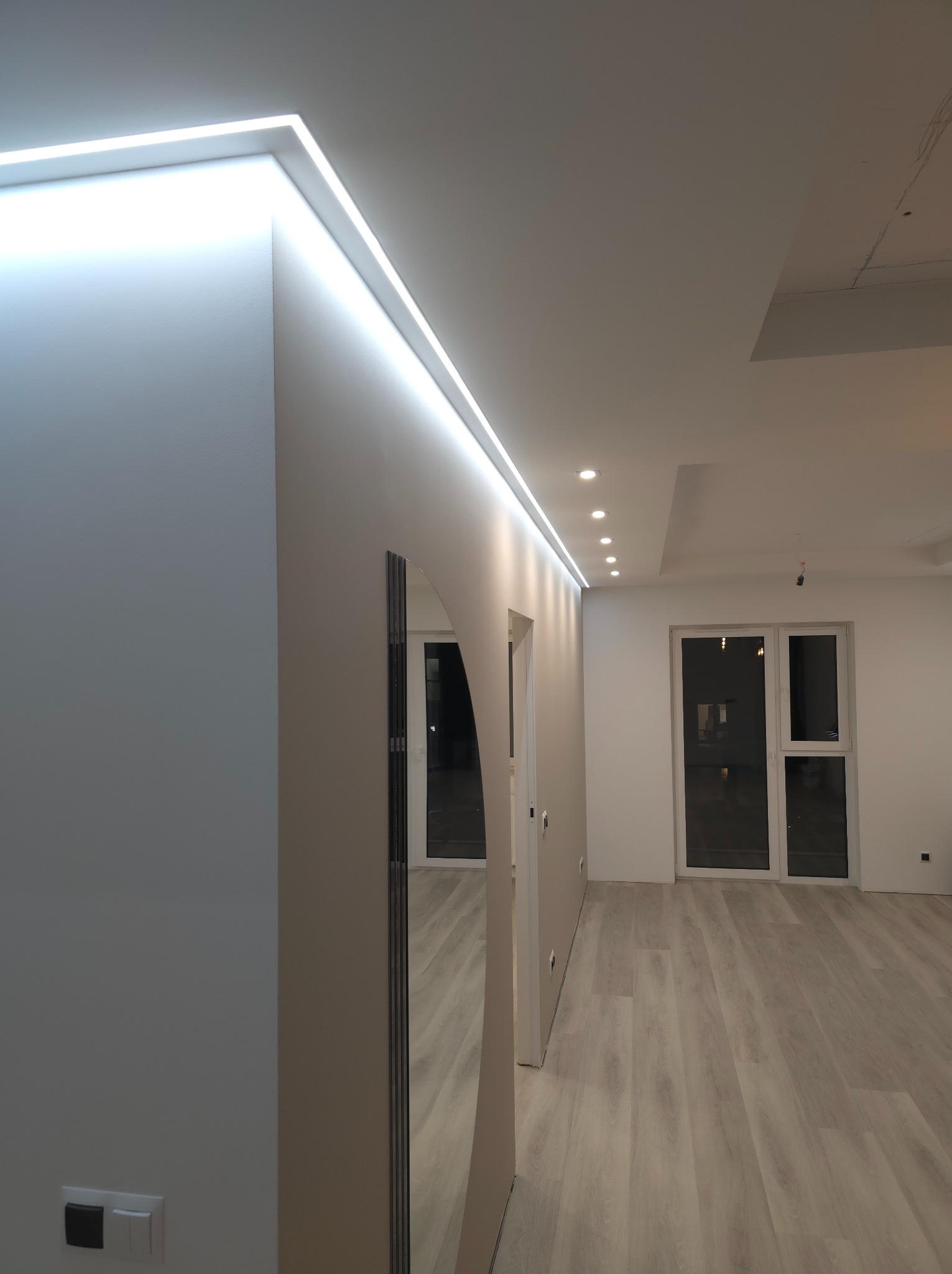 Nowoczesne oświetlenie LED wzdłuż sufitu i ściany, z widocznym parkietem i minimalistycznym wnętrzem w jasnych barwach.