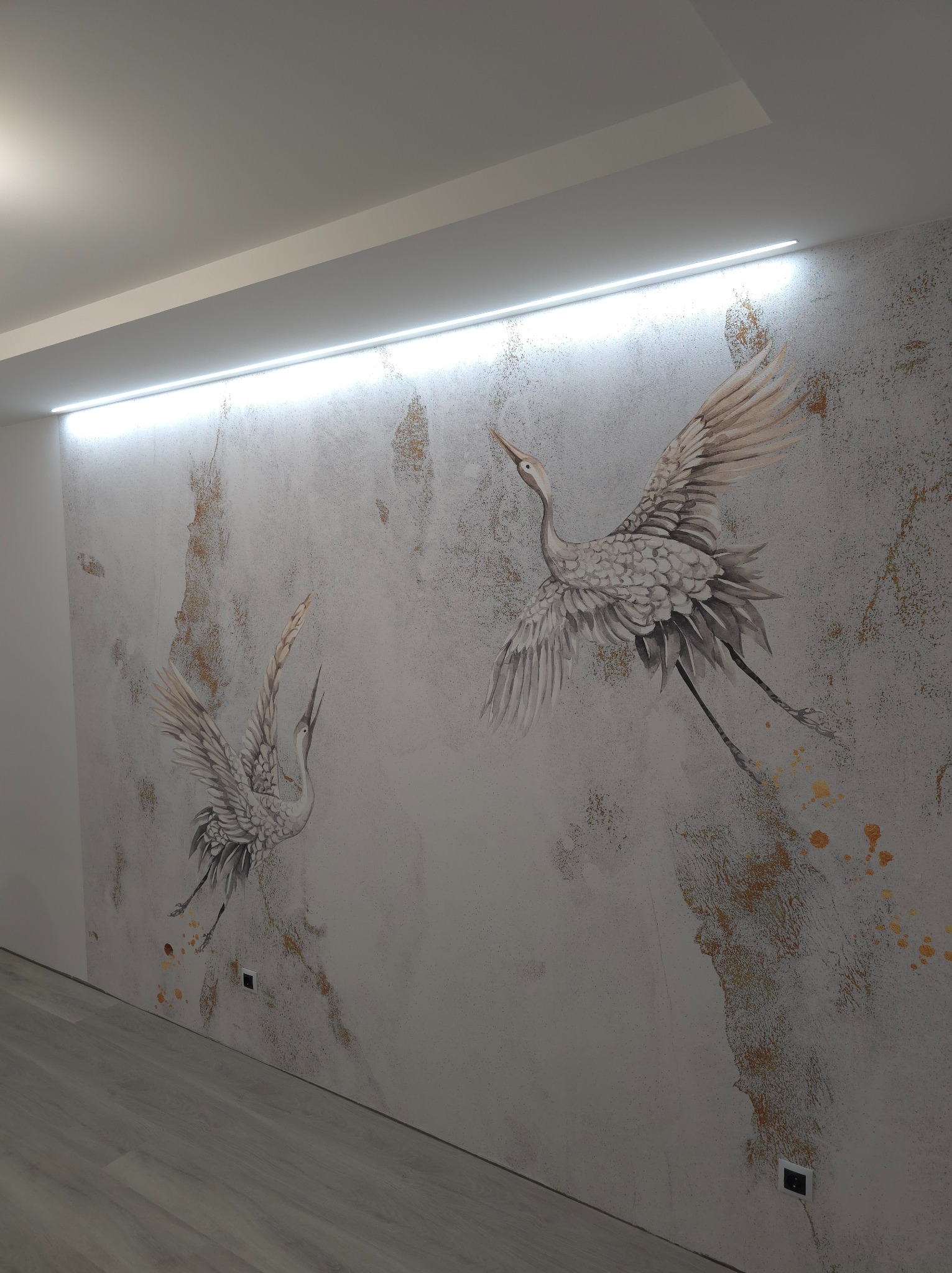 Ściana w pokoju z dekoracyjną tapetą przedstawiającą dwa żurawie w locie na tle szarego betonu z elementami złota, oświetlona listwą LED podsufitową.