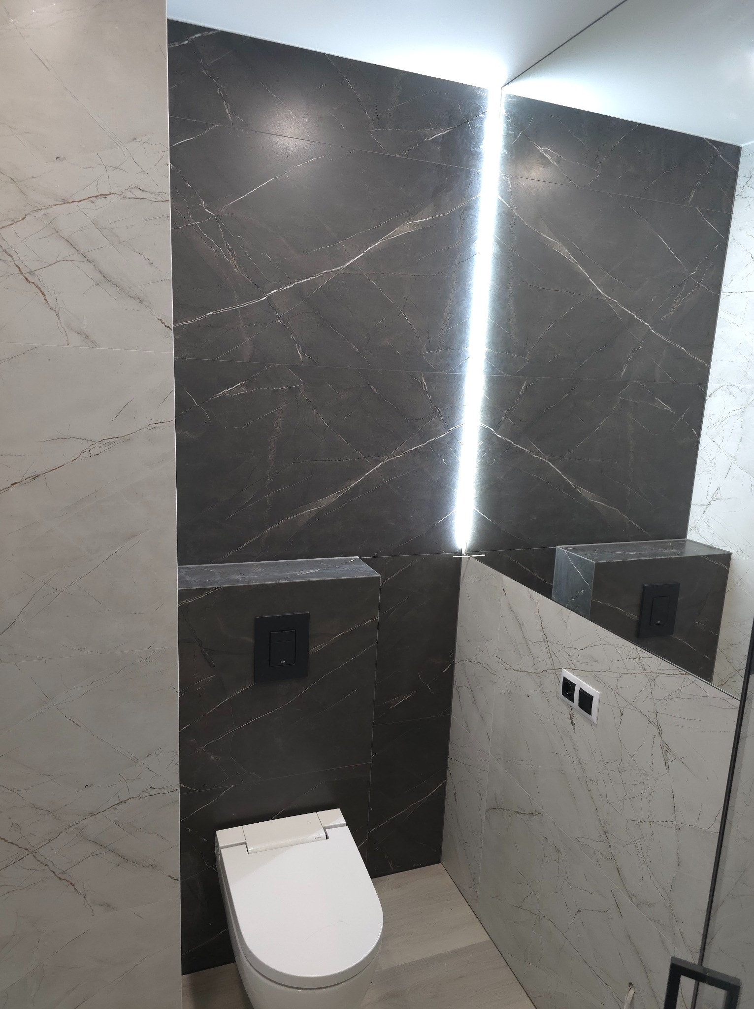 Nowoczesna łazienka z marmurowymi płytkami w odcieniach szarości i bieli, podświetlona pionowym pasem światła, wisząca toaleta z czarnym przyciskiem.