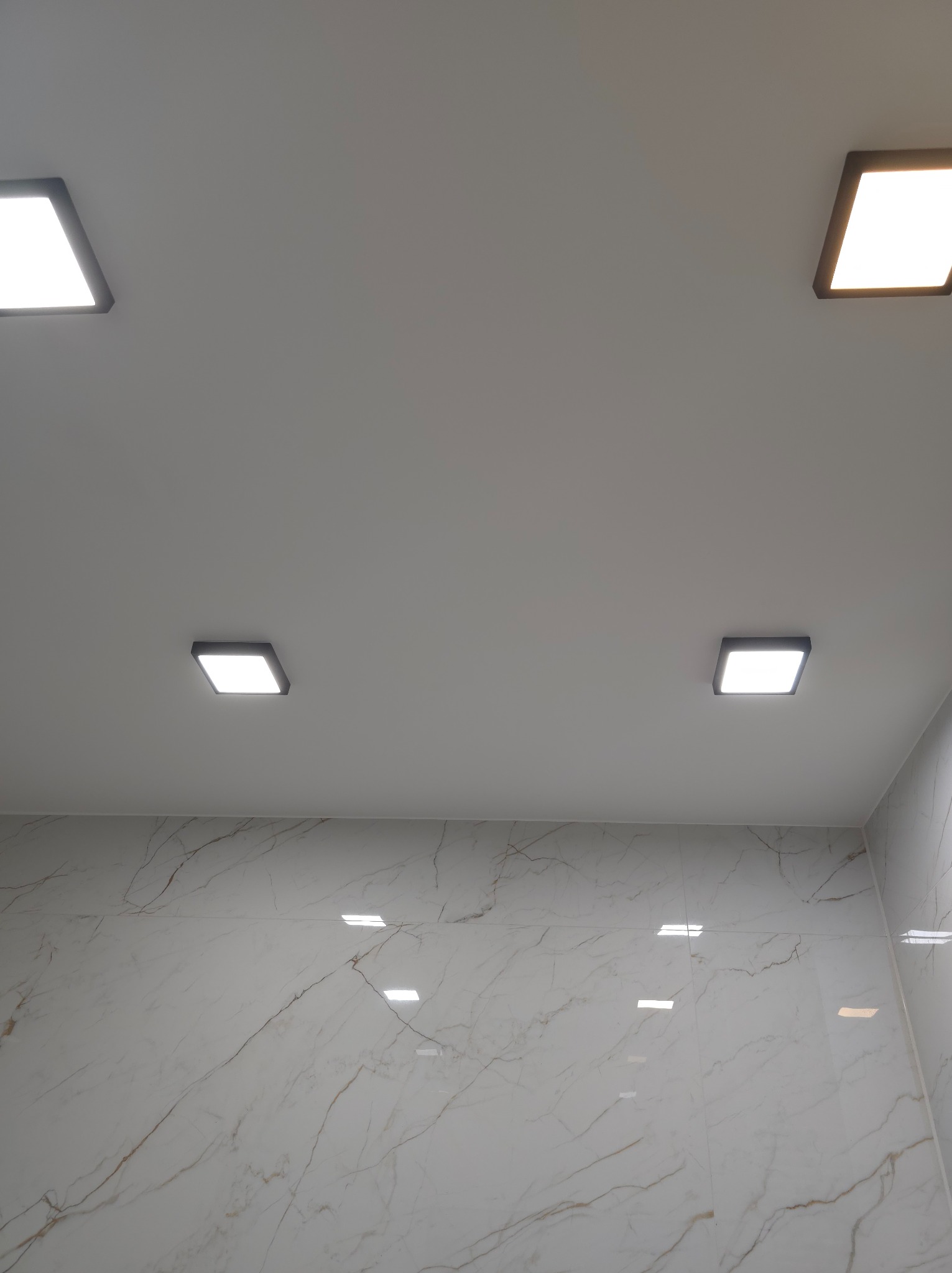 Wykończona łazienka z białymi marmurkowymi płytkami na ścianach i suficie z czterema kwadratowymi lampami LED.