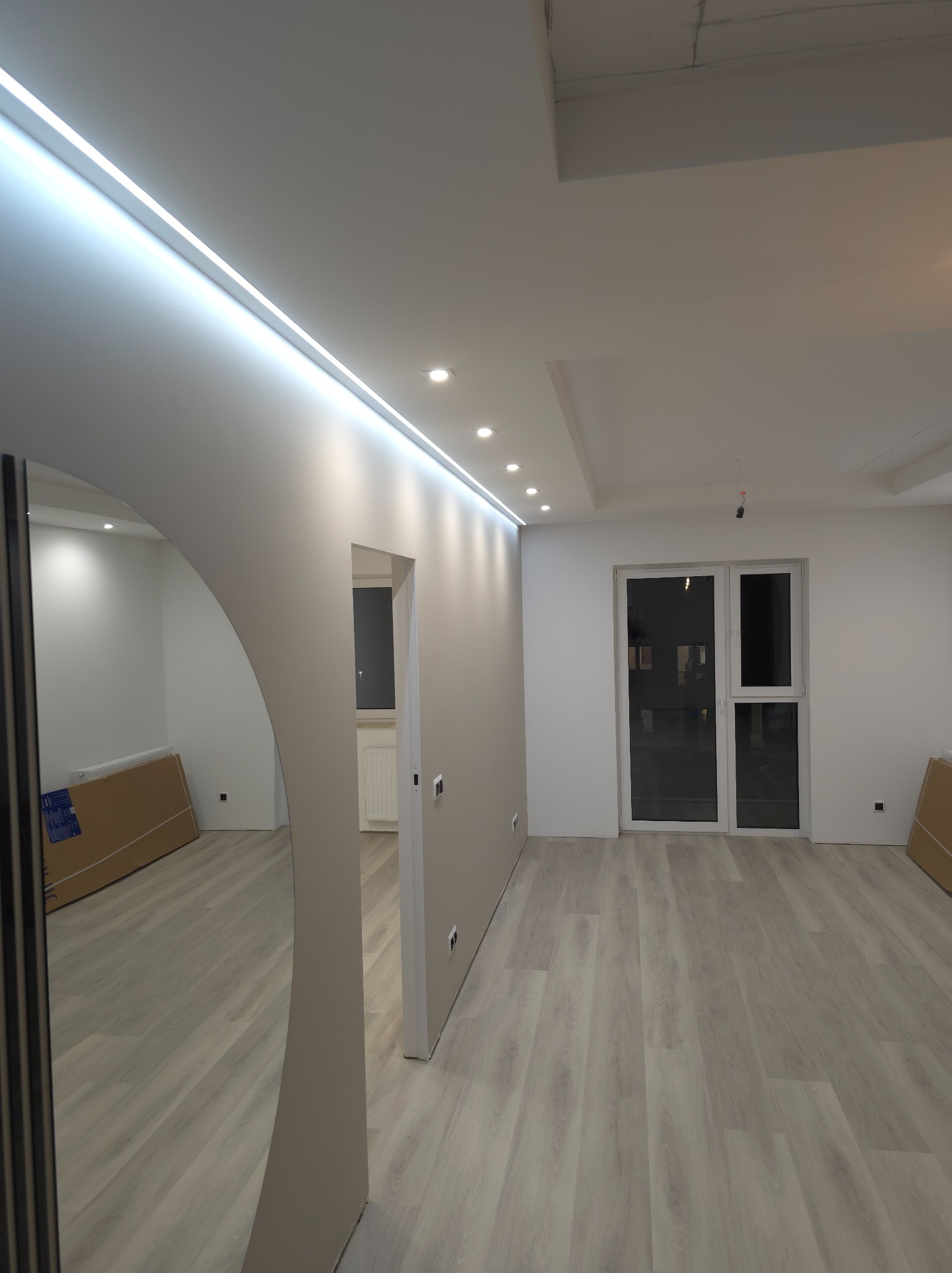 Nowoczesne wnętrze po remoncie: jasne panele podłogowe, ściany pomalowane na szaro, listwa LED w suficie, okrągłe lustro i widok na kolejne pomieszczenie z grzejnikiem.