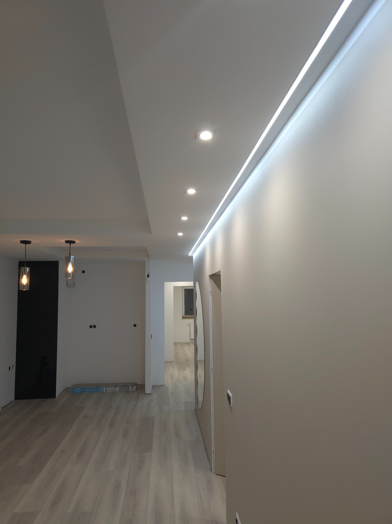 Wnętrze po remoncie: gładkie ściany w odcieniach beżu, podwieszany sufit z oświetleniem LED liniowym i punktowym, panele podłogowe imitujące drewno, dekoracyjne lampy wiszące, drzwi wewnętrzne.