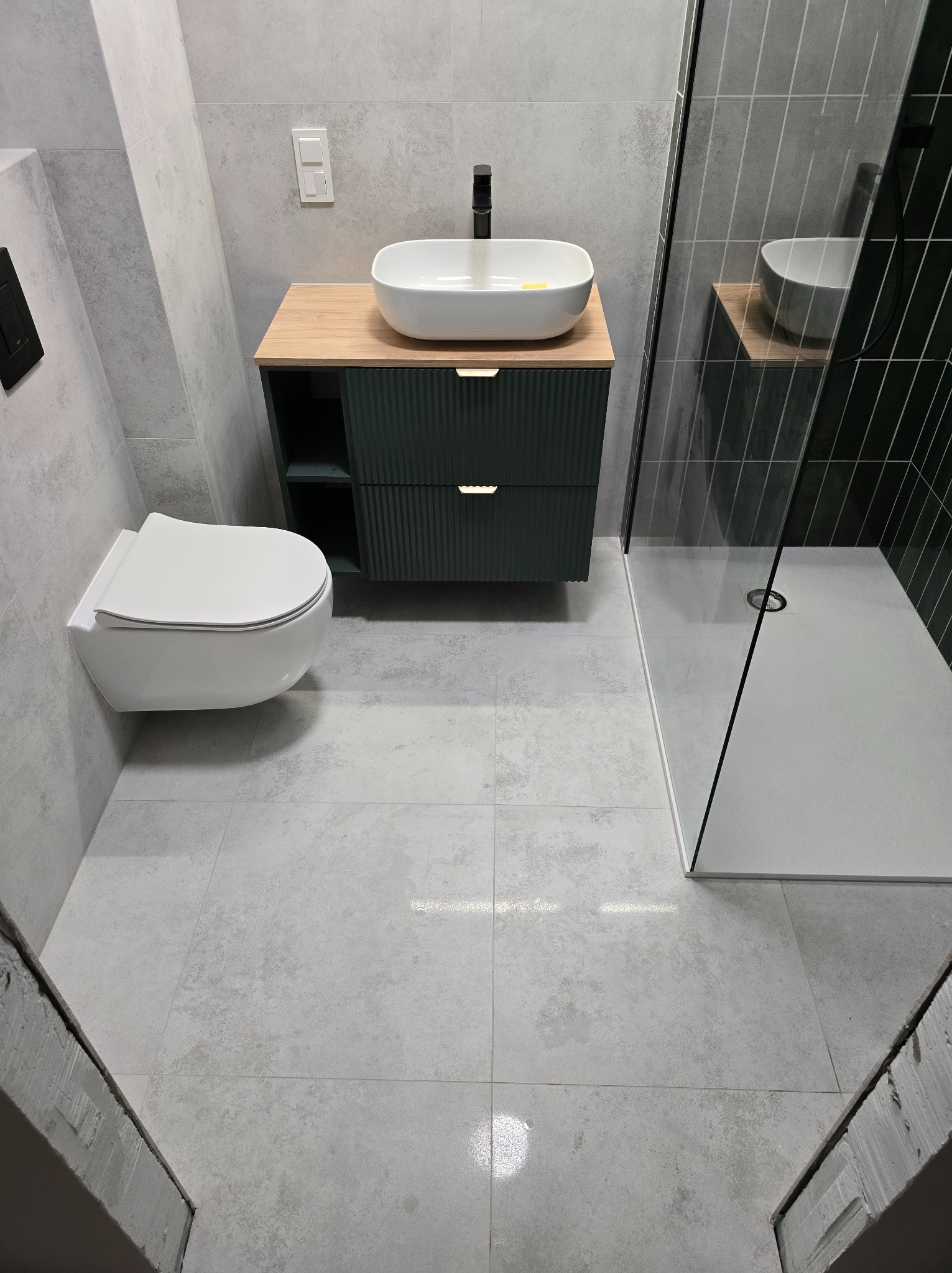 Nowoczesna, szara łazienka z podwieszaną toaletą, szafką umywalkową z ryflowanym frontem i kabiną prysznicową walk-in z czarnymi płytkami na ścianie.