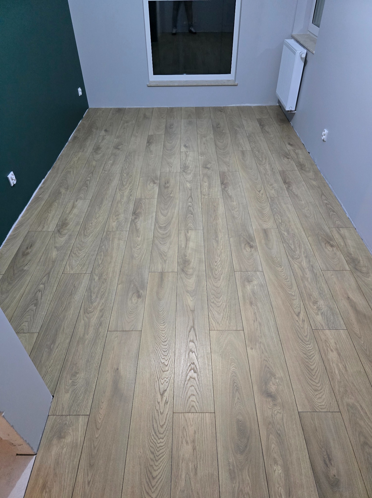 Ułożone panele podłogowe w pokoju z ciemnozieloną i szarą ścianą, widoczny biały grzejnik i okno.