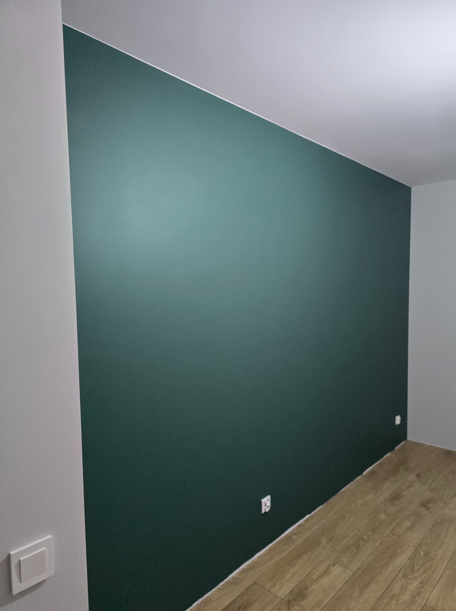 Pomieszczenie po remoncie: ściana pomalowana na ciemnozielony kolor, jasna podłoga z paneli, widoczne gniazdka elektryczne i przełącznik światła.