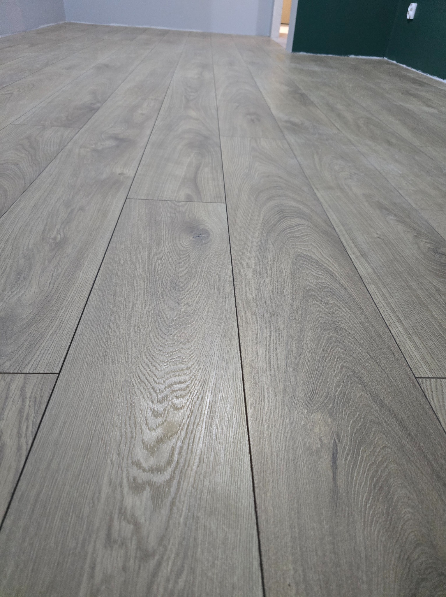 Nowo ułożona, szara podłoga z paneli laminowanych, widok z niskiej perspektywy, z widocznym fragmentem ściany pomalowanej na szaro i zielono.