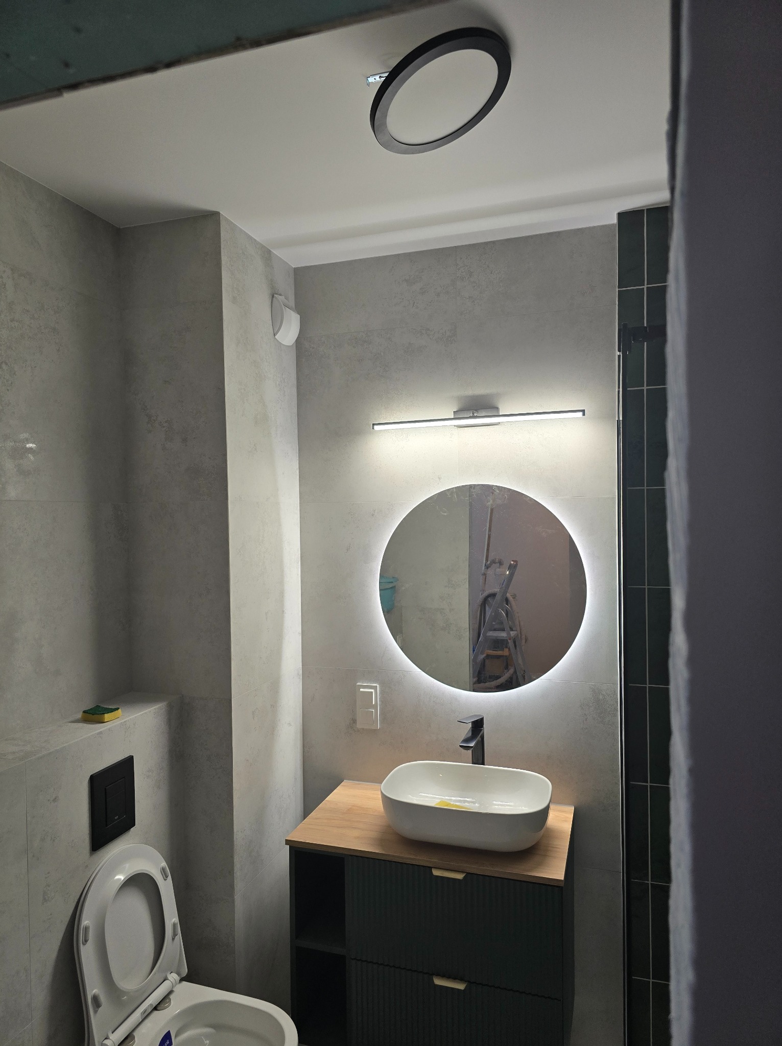Nowoczesna łazienka w szarościach z okrągłym lustrem z podświetleniem LED, drewnianym blatem pod umywalką i toaletą w tle; w odbiciu lustra widoczna drabina malarska.