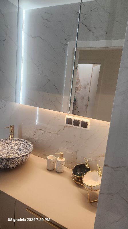 Wykończona łazienka z marmurowymi płytkami, umywalką w stylu vintage z niebieskim wzorem, podświetlanym lustrem i akcesoriami łazienkowymi w odcieniach bieli i złota.