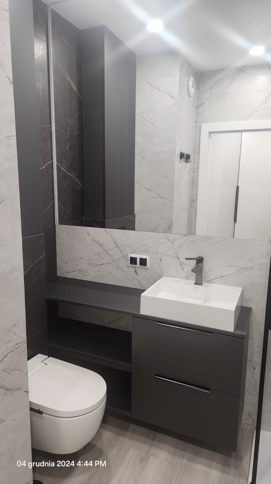 Nowoczesna łazienka z marmurowymi płytkami, wiszącą toaletą i szafką pod umywalkę w kolorze antracytowym, minimalistyczna bateria umywalkowa.