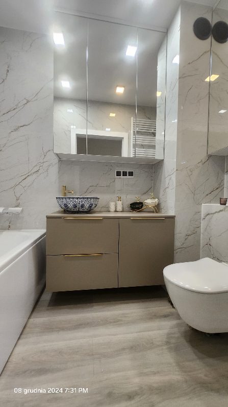 Nowoczesna łazienka z marmurowymi płytkami, beżową szafką pod umywalką z złotymi uchwytami, dekoracyjną umywalką w kolorze niebiesko-białym oraz wiszącą toaletą.