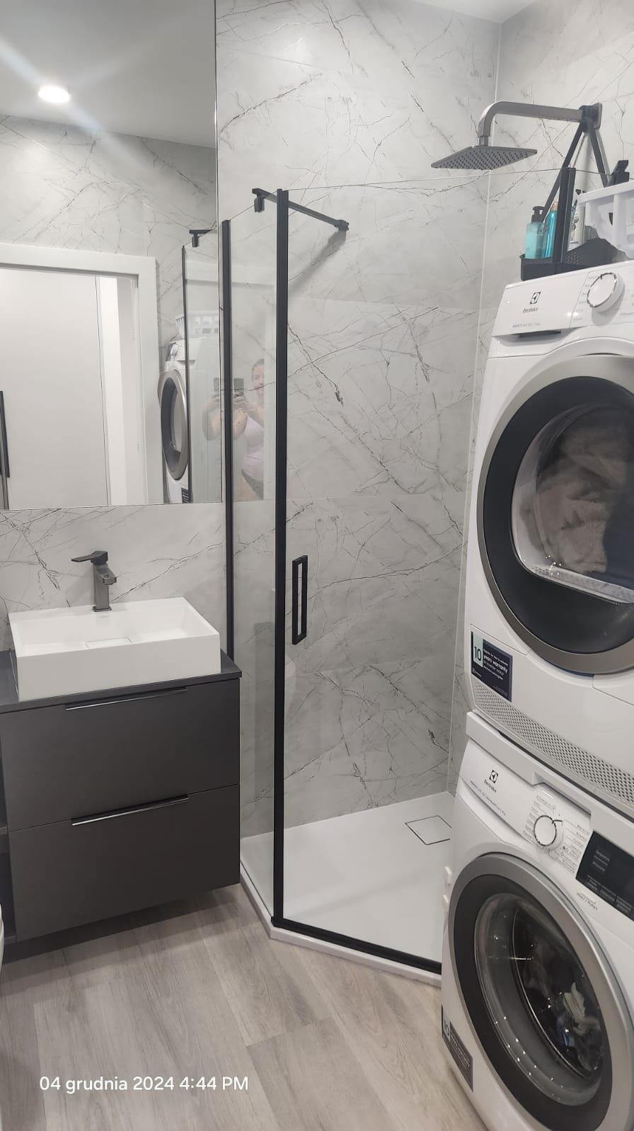 Nowoczesna łazienka z kabiną prysznicową z czarnymi ramami, umywalką nablatową na szarej szafce, pralką i suszarką Electrolux w kolumnie, wykończona płytkami imitującymi marmur.