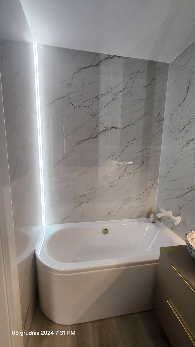 Nowoczesna łazienka z białą wanną, marmurowymi płytkami ściennymi i subtelnym podświetleniem LED w narożniku, uzupełniona minimalistyczną armaturą i drewnopodobną podłogą.