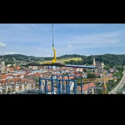 Żuraw podnosi element konstrukcyjny na tle panoramy miasta z kościołem i wzgórzami.