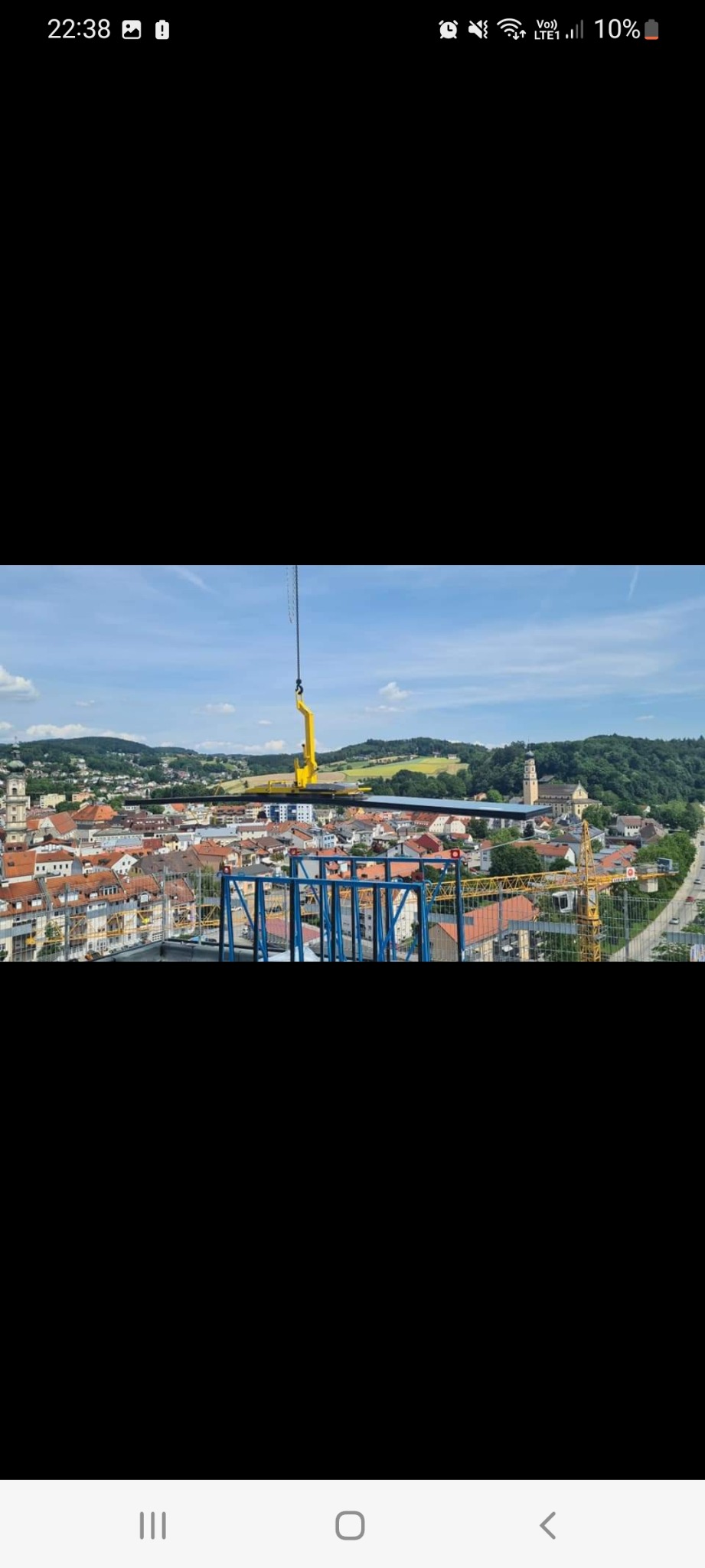 Żuraw podnosi element konstrukcyjny na tle panoramy miasta z kościołem i wzgórzami.