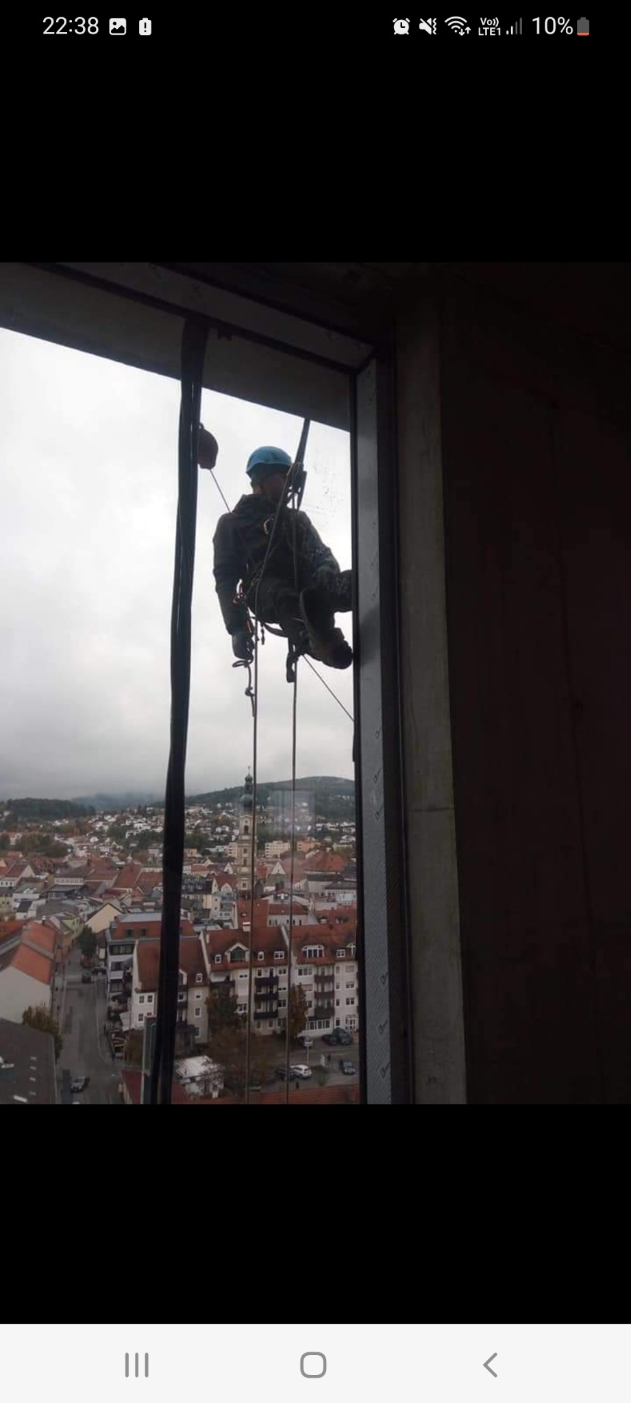 Pracownik wysokościowy w uprzęży i kasku, zawieszony na linach przed oknem podczas pracy, z widokiem na miasto w tle.